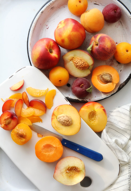 An assortment of stone fruits including peaches, apricots, plums, and nectarines in a bowl and on a cutting board. Some fruits are whole and others have been halved with or without the pit. 