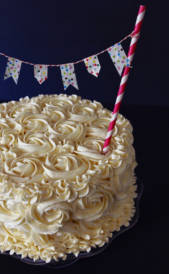 The finished cake covered in white frosting roses with a decretive straw holding up a banner sticking out of the cake. 