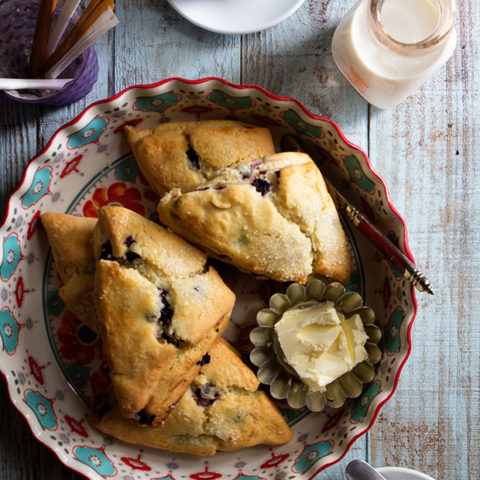 Blueberry scones on a plate with a some mugs on a table.