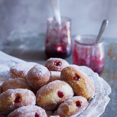 Homemade yeast doughnuts filled with raspberry jam in a bowl next to a jar of raspberry jam.
