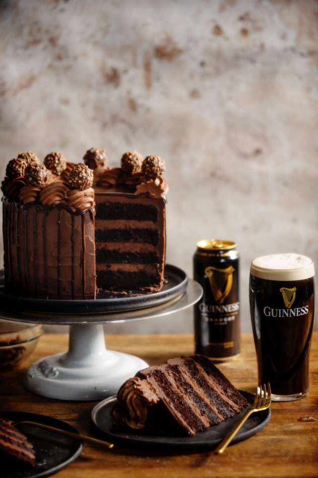 Chocolate Guinness Cake on a cake stand with one slice cut and on a plate. Behind are an empty can of Guinness and a glass full of Guinness beer.