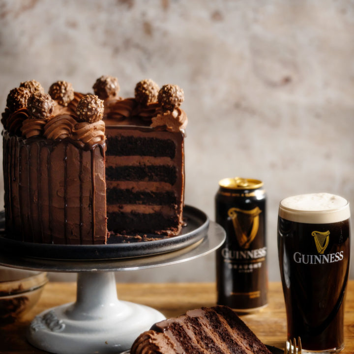 Chocolate Guinness Cake on a cake stand with one slice cut and on a plate. Behind are an empty can of Guinness and a glass full of Guinness beer.